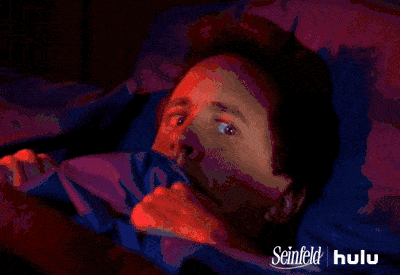 A person lying in bed looks scared, pulling the sheets up to their chin. The image is tinted with red and purple lighting, reminiscent of a school culture's eerie influence. Seinfeld and Hulu logos are visible in the corner.
