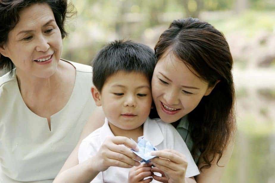 A woman and a child, smiling despite the recent school closures, admire an origami figure together. An older woman joins in their joy, observing warmly. They are outdoors, surrounded by lush greenery.