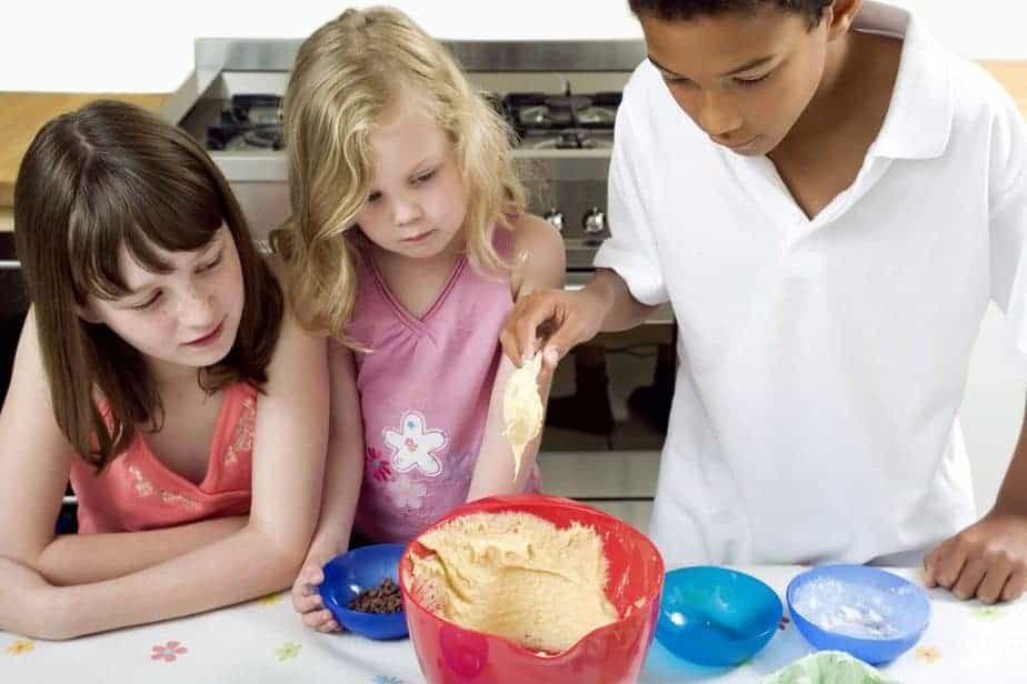 Three children stand around a kitchen table, taking advantage of school closures to bake together. They observe as one scoops batter from a red bowl with a spoon, surrounded by smaller bowls of ingredients. A stove is visible in the background, ready for their culinary creations.