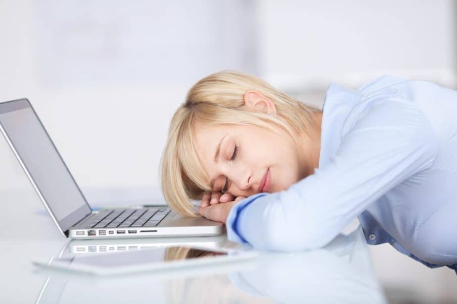 A person with short blonde hair is sleeping on their arms beside a laptop, seemingly exhausted from brainstorming lesson plan ideas. They're wearing a light blue shirt in the bright office or study space.