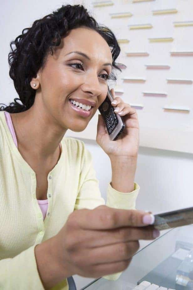 A woman smiles while discussing lesson plan ideas on a cordless phone. She is wearing a light yellow sweater and holding a credit card. In the background, there's an abstract art piece on the wall with colorful lines.