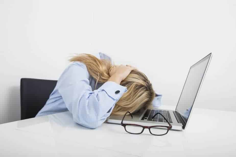 A person, likely searching for lesson plan ideas, sits at a desk with their head resting on a laptop keyboard, hands holding their head in frustration or fatigue. Glasses rest on the table beside them, and they wear a light blue shirt.