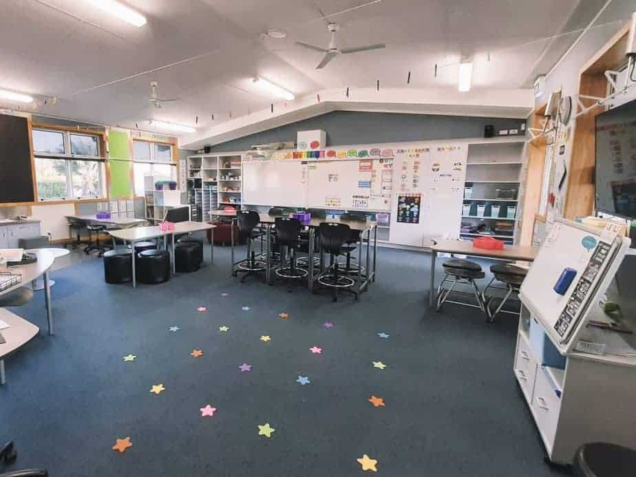 A brightly lit classroom with student tables and stools, a whiteboard, and educational posters on the walls. The floor is decorated with colorful star-shaped stickers. Shelves along the sides hold books and supplies.