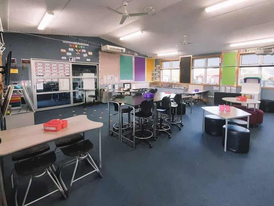 A spacious classroom with several tables and chairs. The room has colorful posters on the walls, large windows, and a digital board. Light streams in through the windows, creating a bright and inviting environment. Boxes and supplies are organized neatly.