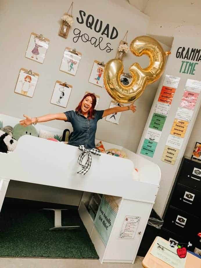 A person with red hair happily spreads their arms in a classroom setting. Behind them, a large gold balloon shaped like the number 3 is visible. The walls are decorated with the phrases Squad Goals and Grammar Time alongside student artwork.