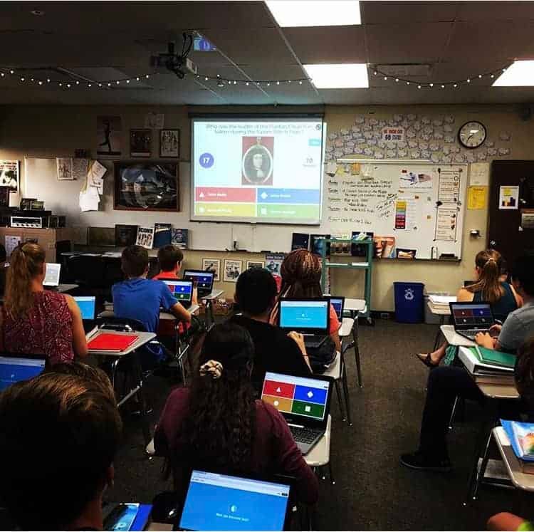 A classroom with students sitting at desks using laptops, looking at a projected screen displaying a quiz game. The room is decorated with string lights and posters, and a whiteboard is visible at the front.