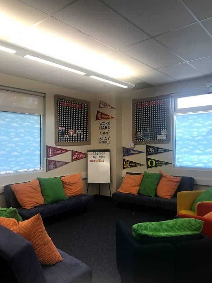 A cozy room with dark sofas and colorful cushions arranged around the walls. Bright posters and college pennants decorate the walls, along with a motivational sign that reads, Work Hard, Stay Humble. Soft natural light from the windows illuminates the space.