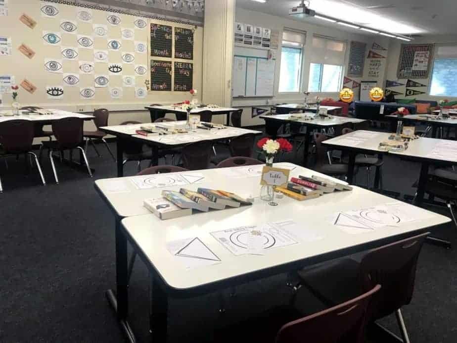A well-organized classroom with white tables arranged in groups, each displaying papers, books, and stationary trays. Walls are decorated with educational posters and pennants. Vases with flowers are placed on the tables.