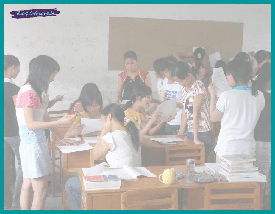 A group of students in a classroom, navigating challenges together around desks, engaged with papers. Some are standing, while others are seated, discussing and sharing documents. The room features books and a large brown bulletin board on the wall.