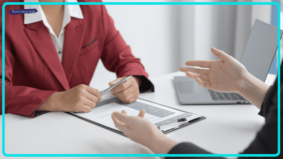 A person in a red blazer holding a clipboard with a resume sits across from another person gesturing with their hands. A laptop is open on the table, perhaps ready to dive into teacher interview questions. The setting appears to be a professional interview or meeting.
