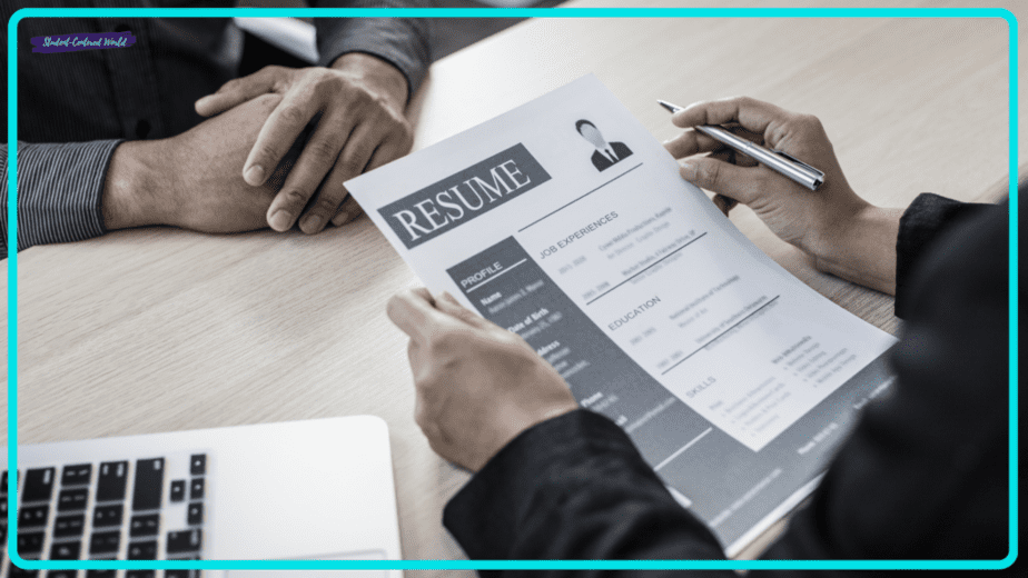 A person holds a resume while sitting at a table, discussing teacher interview questions with another individual. A laptop is open on the table, and both are dressed in business attire, indicating this is likely a professional job interview setting.