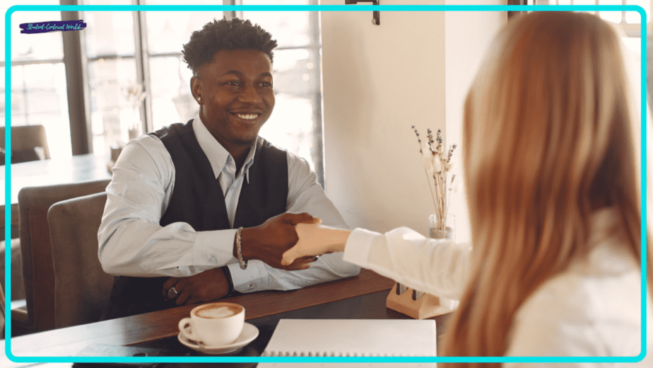 A man and woman are sitting at a table in a well-lit cafe, smiling and shaking hands. A notebook filled with teacher interview questions and a coffee cup sit on the table, suggesting a friendly meeting or an insightful interview.