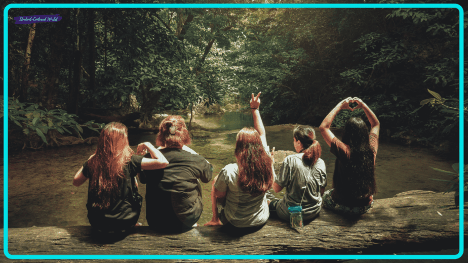 Five people with long hair sit on a log beside a creek, surrounded by lush forest. Facing away from the camera, some make peace and heart signs with their hands, inspiring thoughts on how to motivate students. Sunlight filters through the trees, creating a serene atmosphere.