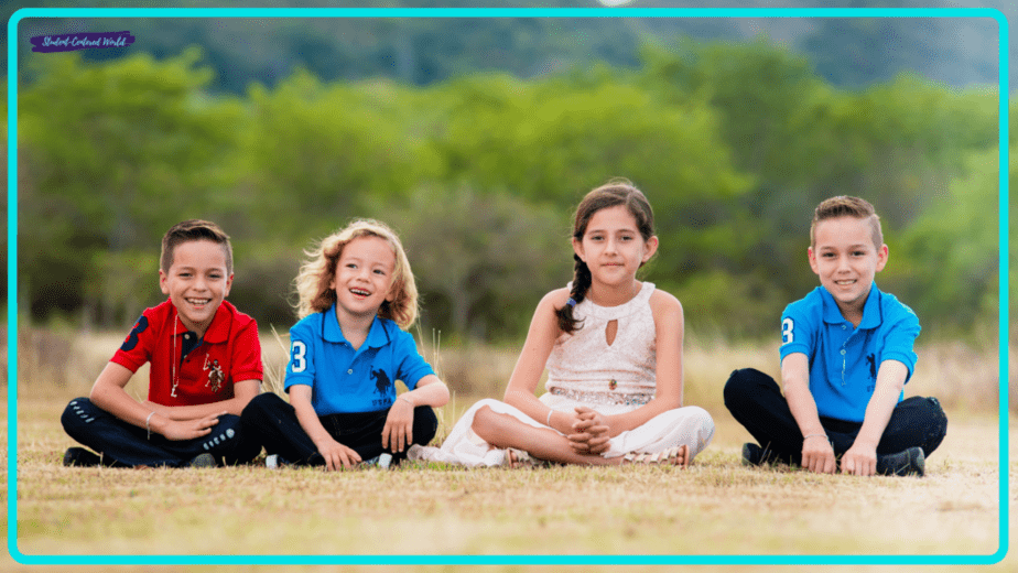 Four children are sitting on the grass in a park, perhaps sparking ideas for how to motivate students with nature's inspiration. Two boys in red and blue shirts sit on the left, a girl in a white dress is in the middle, and another boy in blue sits on the right, framed by trees in the background.