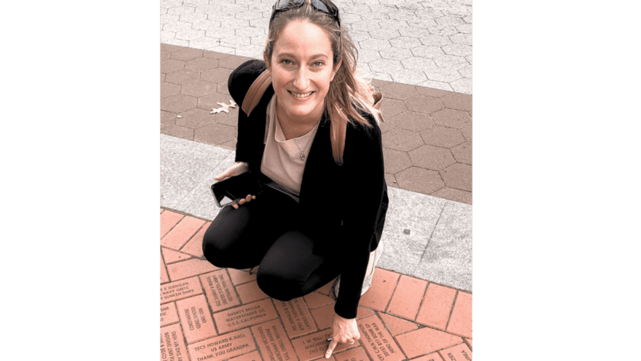 A woman smiling and kneeling on a brick path, pointing at an engraved brick. She holds a smartphone in her other hand. The path consists of engraved bricks forming a pattern. She wears a black jacket and sunglasses on her head.