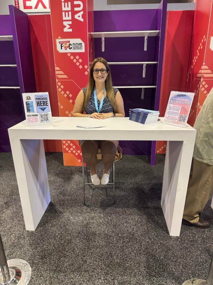 A woman sits at a white table in a conference setting with shelves and banners in the background. She wears glasses and a patterned dress. QR codes and brochures are on the table.