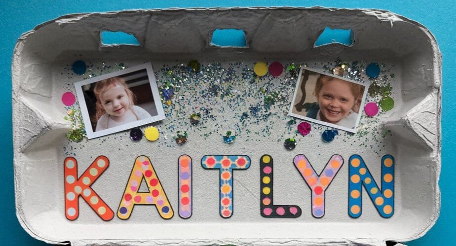 A decorated egg carton with Kaitlyn spelled out in colorful, patterned letters. Two photos of a smiling child are above the name, surrounded by glitter, sequins, and colorful dots on a blue background.