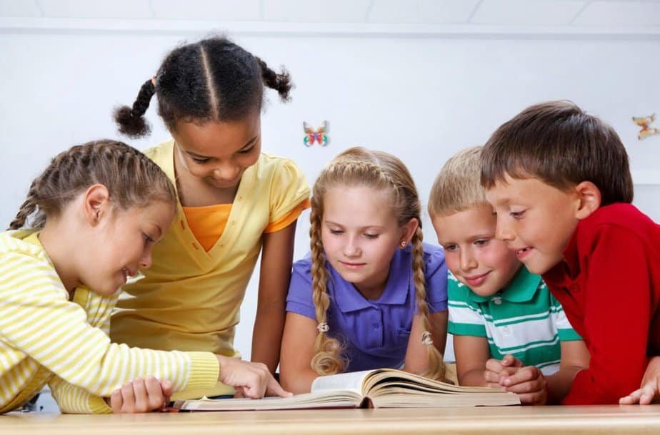 Five children are gathered around a book on a table, engaged in reading. They are smiling and dressed in colorful shirts. The setting appears to be a classroom, with butterfly decorations on the wall in the background.