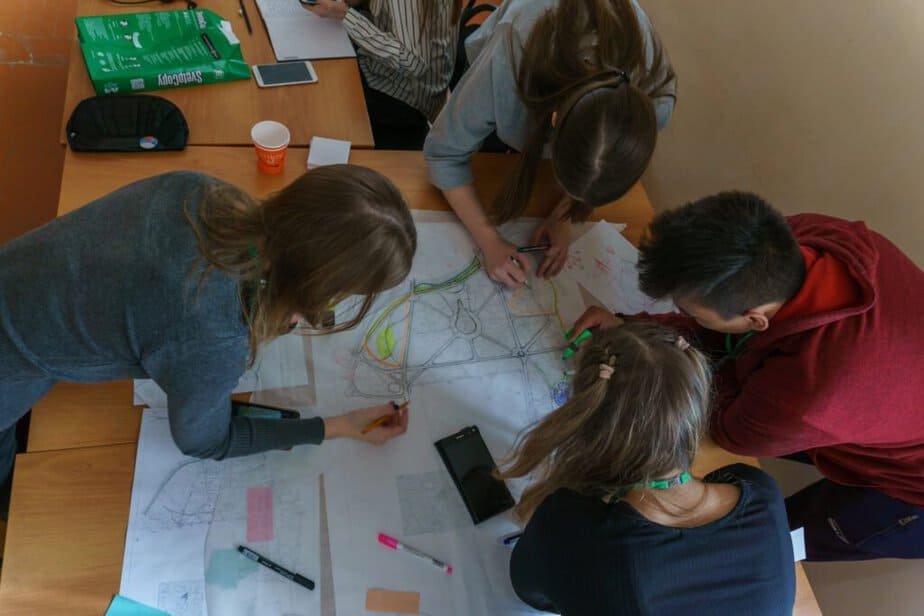 A group of people are gathered around a table covered with large maps and papers. They are actively discussing and drawing on the maps with colored pencils. Various stationery items and a paper cup are scattered on the table. student centered learning examples