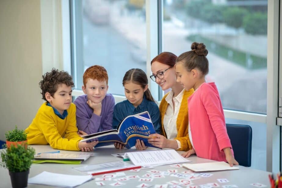 A teacher and four children are gathered around a table, with Student Engagement Activities with a book titled English Grammar. The children appear focused, while the teacher smiles, offering guidance. The setting is a bright classroom with large windows in the background.