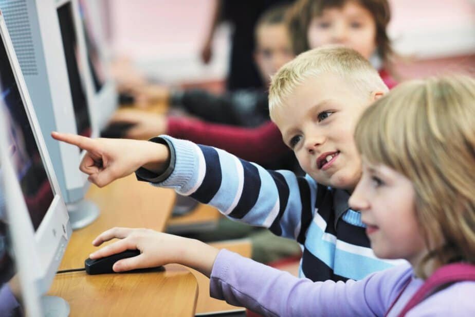 Children sitting at a row of computers, focused on the screens. A boy points at one screen, while a girl next to him looks on attentively. Other children are visible in the background, also engaged with computers.