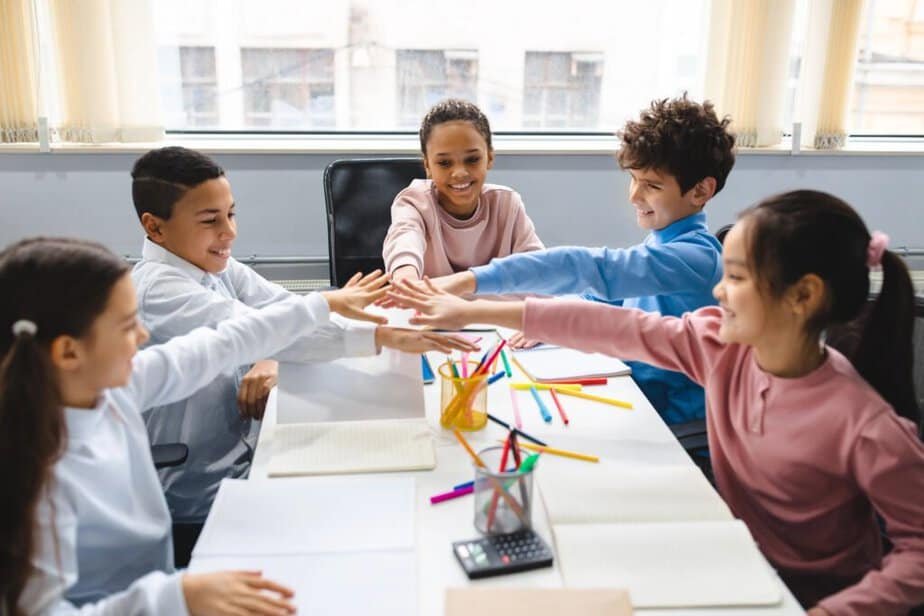 Five children sit around a classroom table, smiling and stacking their hands in the center. They are surrounded by open notebooks and colorful pencils. The background shows large windows letting in natural light.