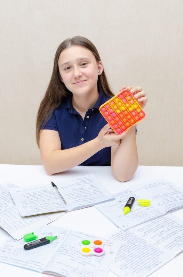 A young girl in a navy shirt holds a colorful pop-it toy. She is sitting at a table with open notebooks, highlighting markers, and a pen. She smiles gently against a plain background.