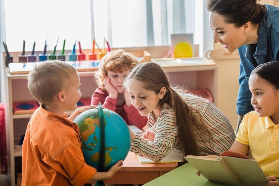 Children gathered around a table in a classroom. A boy in an orange shirt holds a globe, while others, including a girl leaning in and a teacher smiling, look on. Art supplies are visible in the background. Open-Ended Prompts for Student Behavior