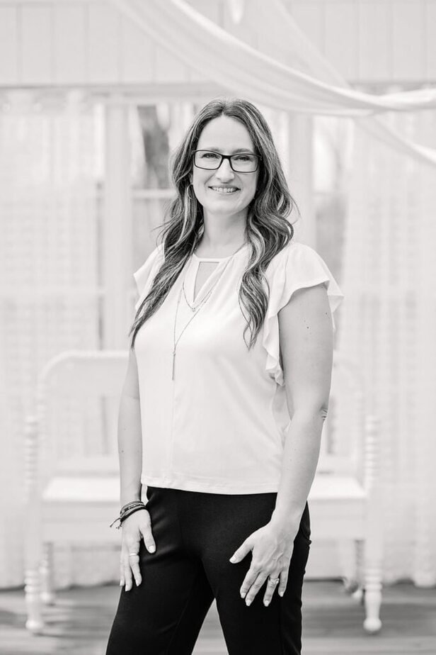 A woman with long, wavy hair and glasses is smiling, standing indoors. She wears a light blouse with flutter sleeves and dark pants, accessorized with a long necklace. The background is softly lit and neutral.