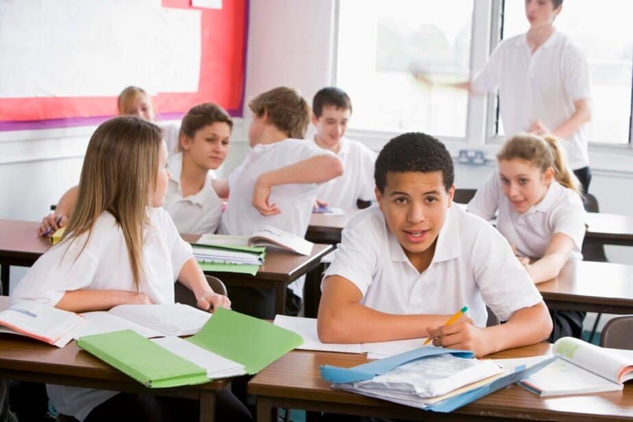 A classroom with several students in white uniforms. One boy sits at a desk in the foreground, looking at the camera, while others talk and interact in the background. Open books and folders are on the desks. How to Deal with Disruptive Students: Teacher Guide