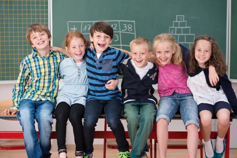 Six smiling children sit close together on a bench in a classroom, arms around each other. They are in front of green chalkboards with drawings and math problems. The atmosphere is cheerful and friendly.