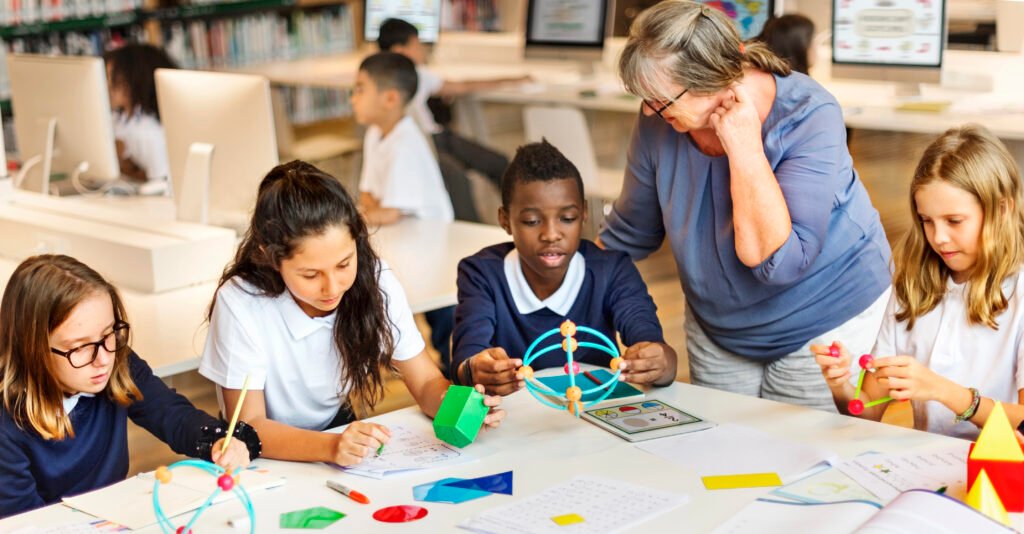 A teacher assists a diverse group of elementary students working with colorful geometric shapes at a table in a classroom with computers and bookshelves in the background.