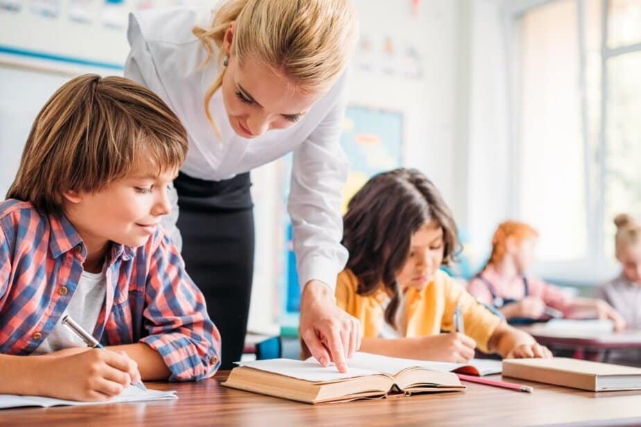 A teacher leans over a desk to help a young student with his work in a classroom, while other students write in their notebooks in the background, sehlping with Strategies for Setting Boundaries with Students and Parents