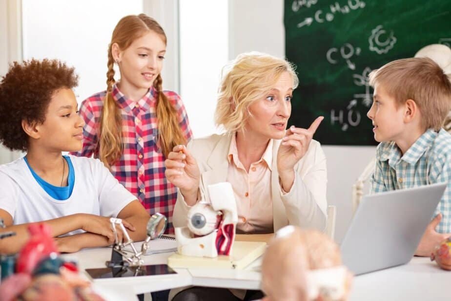 A teacher explains a science concept to three attentive students in a classroom, using anatomical models and a laptop, with a chalkboard visible in the background.