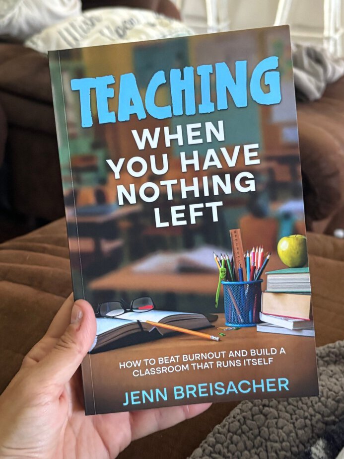 A hand holds the book Teaching When You Have Nothing Left by Jenn Breisacher—a must-read for those feeling burnt out before school even begins. The cover features a cluttered teacher’s desk filled with supplies, books, and an apple.