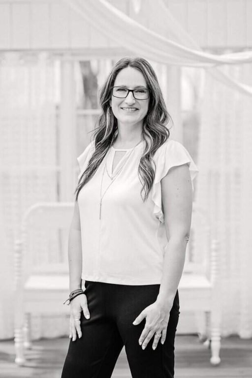 A woman with long, wavy hair and glasses is smiling, standing indoors. She wears a light blouse with flutter sleeves and dark pants, accessorized with a long necklace. The background is softly lit and neutral.